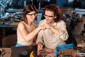 Young man and woman in protective glasses doing experiments in robotics in a laboratory using a computer. Giving each other a cam, robot and tools on the table